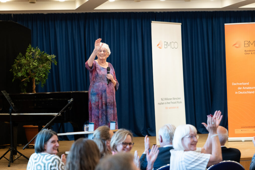 Lore Auerbach winkt den musizierenden Kindern im Saal, das Publikum applaudiert zum Dank für die Musik.
